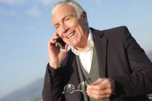 Retired age man on a cell phone call outside with blue sky. Photo compliments of Andrea Piacquadio via Pexels.com